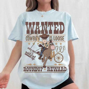 Pode incluir: T-shirt azul claro com um cowboy de desenho animado montado a cavalo e o texto castanho "WANTED COWBOY ON THE LOOSE". O design inclui estrelas, um la&ccedil;o e as palavras "GIVE A HOLLER & SADDLE UP!" e "ROUNDUP REWARD".