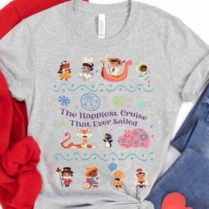 May include: A light gray t-shirt featuring a colorful graphic with the text "The Happiest Cruise That Ever Sailed." The design includes cartoon characters, flowers, and a boat. The shirt is paired with blue jeans and a red sweatshirt.
