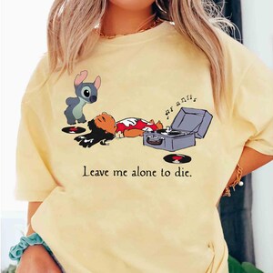 May include: Yellow t-shirt featuring a cartoon illustration of Lilo and Stitch with a record player. The image includes the text "Leave me alone to die."