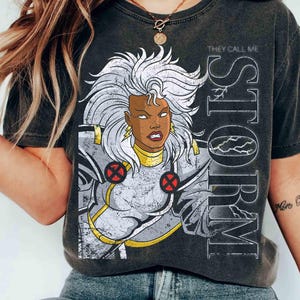 Könnte beinhalten: Ein anthrazitfarbenes T-Shirt mit einem Grafikdruck der Figur Storm mit weißem Haar, die ein silbernes und goldenes Kostüm mit roten X-Men-Emblemen trägt. Der Text "THEY CALL ME STORM" ist vertikal in großen weißen Buchstaben rechts aufgedruckt.