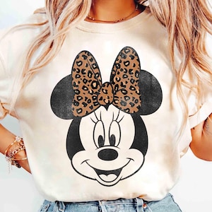 May include: Cream-colored t-shirt featuring a black and white Minnie Mouse graphic. The Minnie Mouse design includes a leopard print bow and black ears. The shirt is a casual, everyday wear item.