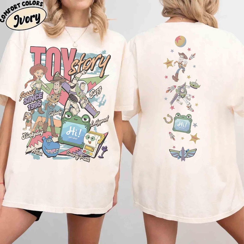 Op de afbeelding: Een ivoorkleurig t-shirt met korte mouwen en afbeeldingen van Toy Story-personages. De voorkant toont Jessie, Woody, Buzz Lightyear, Slinky Dog en andere figuren met de tekst Toy Story, Since 1995 en Hi. De achterkant toont een verticale opstelling van figuren.