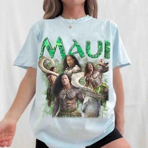 May include: A light blue short-sleeve t-shirt featuring a graphic print of characters from the movie Maui. The design includes the text MAUI in large green letters, images of figures with tribal tattoos, a large fish hook, and a shark.