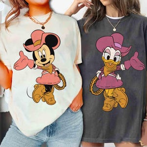 Puede incluir: Dos camisetas con los personajes de dibujos animados Minnie Mouse y Daisy Duck con atuendo de vaquera. Minnie Mouse est&aacute; en una camiseta color crema, y Daisy Duck est&aacute; en una camiseta gris. Ambos personajes visten sombreros, vestidos y botas rosas, con un detalle de cuerda.