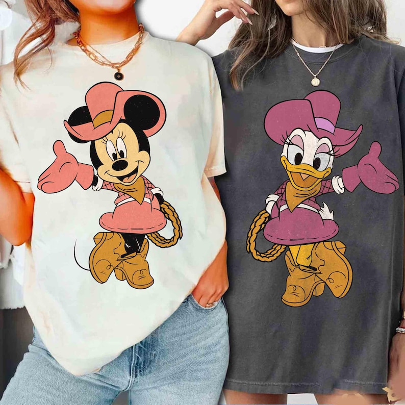 Puede incluir: Dos camisetas con los personajes de dibujos animados Minnie Mouse y Daisy Duck con atuendo de vaquera. Minnie Mouse est&aacute; en una camiseta color crema, y Daisy Duck est&aacute; en una camiseta gris. Ambos personajes visten sombreros, vestidos y botas rosas, con un detalle de cuerda.