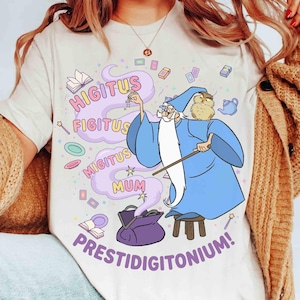 May include: A cream-colored t-shirt featuring a cartoon wizard in a blue robe and hat, holding a wand. The shirt has the words "HIGITUS FIGITUS MIGITUS MUM PRESTIDIGITONIUM!" in a whimsical font, with magical elements.