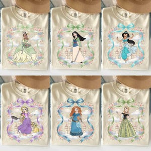 May include: Cream-coloured t-shirts featuring various Disney princesses. Each shirt showcases a princess illustration within a floral and ribbon frame. The princesses include Tiana, Mulan, Jasmine, Rapunzel, Merida, and Elsa.
