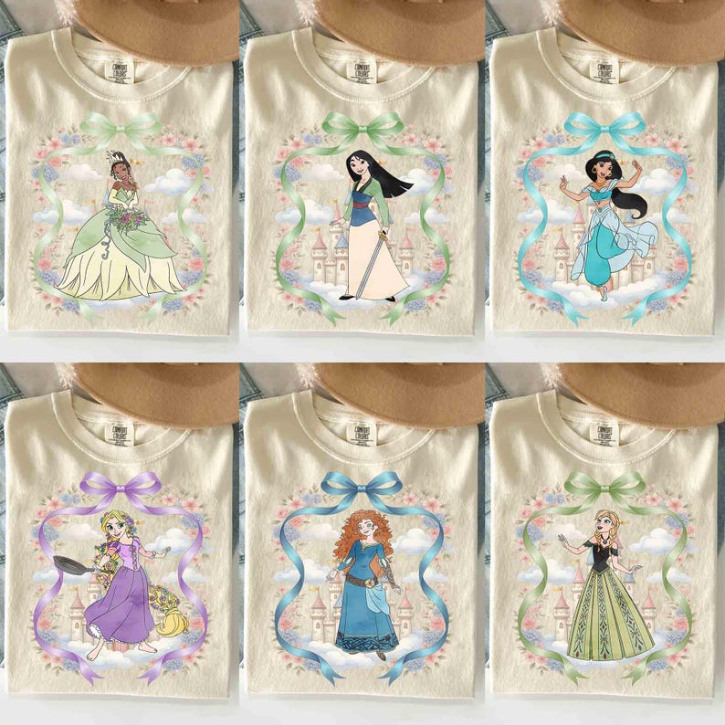 May include: Cream-coloured t-shirts featuring various Disney princesses. Each shirt showcases a princess illustration within a floral and ribbon frame. The princesses include Tiana, Mulan, Jasmine, Rapunzel, Merida, and Elsa.