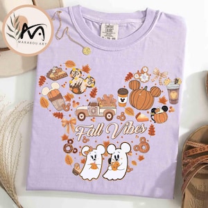 May include: A light purple t-shirt with a fall-themed graphic. The design features a Mickey Mouse-shaped arrangement of pumpkins, fall leaves, and a truck. The words "Fall Vibes" are written in a script font. The shirt is paired with sandals.