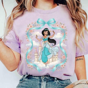 May include: A lilac t-shirt with a cartoon illustration of a princess in a teal outfit, standing in front of a castle. The design includes a teal ribbon, flowers, and clouds. The shirt is made of a soft material.