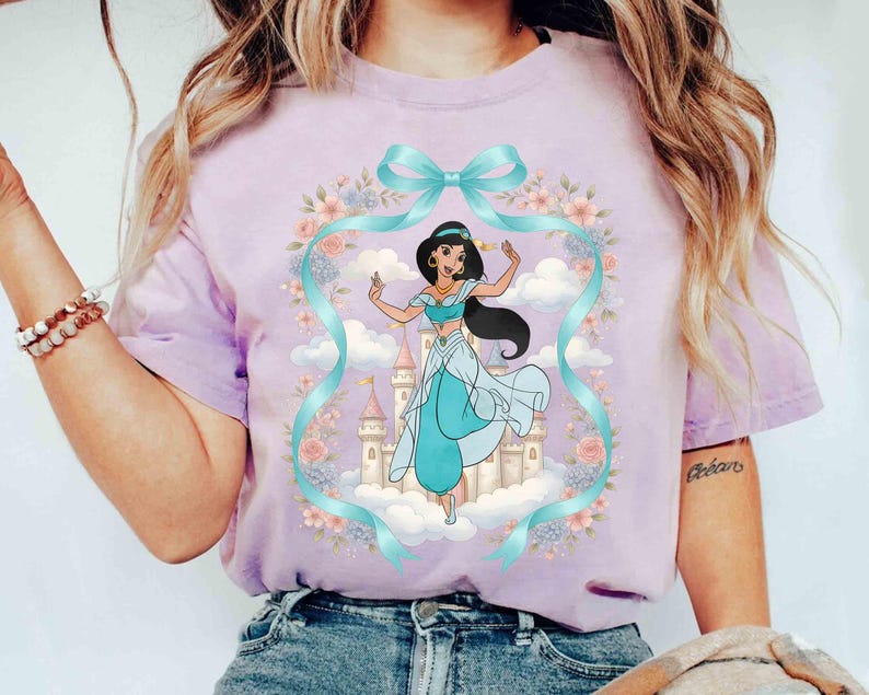 May include: A lilac t-shirt with a cartoon illustration of a princess in a teal outfit, standing in front of a castle. The design includes a teal ribbon, flowers, and clouds. The shirt is made of a soft material.