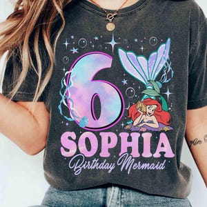 Puede incluir: Camiseta gris oscuro con un dise&ntilde;o de sirena. El dise&ntilde;o incluye el n&uacute;mero 6 en un degradado p&uacute;rpura y azul, el nombre "SOPHIA" y las palabras "Birthday Mermaid". Tambi&eacute;n aparece una sirena de dibujos animados.