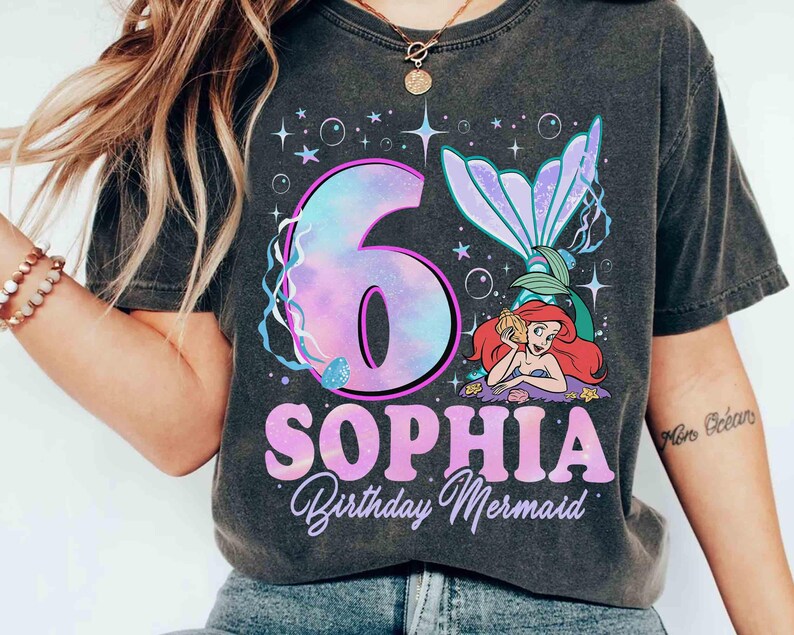 Puede incluir: Camiseta gris oscuro con un dise&ntilde;o de sirena. El dise&ntilde;o incluye el n&uacute;mero 6 en un degradado p&uacute;rpura y azul, el nombre "SOPHIA" y las palabras "Birthday Mermaid". Tambi&eacute;n aparece una sirena de dibujos animados.