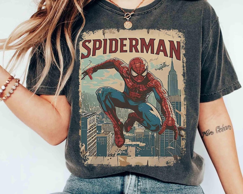 May include: Dark gray t-shirt featuring a vintage-style Spider-Man graphic. The design shows Spider-Man in a dynamic pose against a cityscape backdrop, with the word "SPIDERMAN" in bold letters. The shirt has a worn, distressed look.