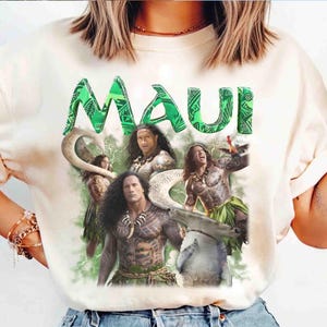 May include: A cream-colored short-sleeve t-shirt featuring a graphic print of the character Maui from the movie Moana. The design includes the green text MAUI at the top, multiple depictions of the character with tattoos, a large fish hook, and a shark.
