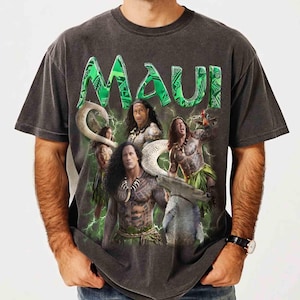 May include: A charcoal gray short-sleeve t-shirt featuring a large graphic print of the character Maui from the movie Moana. The design includes the green text MAUI at the top, multiple depictions of the character, and green lightning bolt accents.