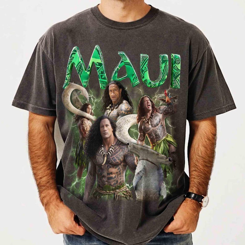 May include: A charcoal gray short-sleeve t-shirt featuring a large graphic print of the character Maui from the movie Moana. The design includes the green text MAUI at the top, multiple depictions of the character, and green lightning bolt accents.