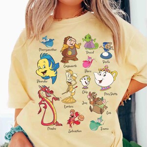 Puede incluir: Una camiseta amarillo claro con personajes de dibujos animados coloridos, incluyendo Flounder, Mushu y la Sra. Potts. Los nombres de los personajes están impresos debajo de cada imagen. La camiseta tiene cuello redondo y mangas cortas.