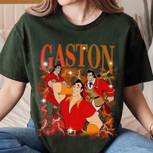 Pu&ograve; includere: T-shirt verde scuro con una grafica di Gaston de La Bella e la Bestia. Il design include la parola "GASTON" in arancione, con illustrazioni di Gaston in varie pose, su uno sfondo di fulmini.