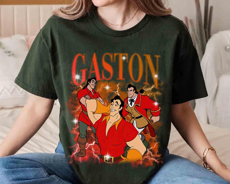 Pu&ograve; includere: T-shirt verde scuro con una grafica di Gaston de La Bella e la Bestia. Il design include la parola "GASTON" in arancione, con illustrazioni di Gaston in varie pose, su uno sfondo di fulmini.