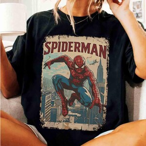 May include: Black t-shirt featuring a vintage-style Spider-Man graphic. The design includes the word "SPIDERMAN" in red, with an illustration of the superhero in action against a cityscape backdrop. The shirt is a classic crew neck.