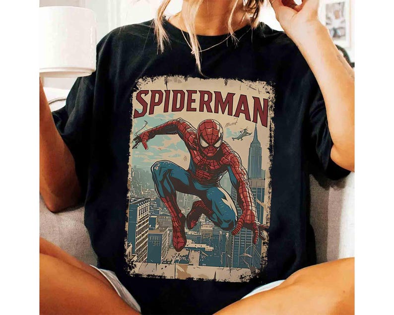 May include: Black t-shirt featuring a vintage-style Spider-Man graphic. The design includes the word "SPIDERMAN" in red, with an illustration of the superhero in action against a cityscape backdrop. The shirt is a classic crew neck.