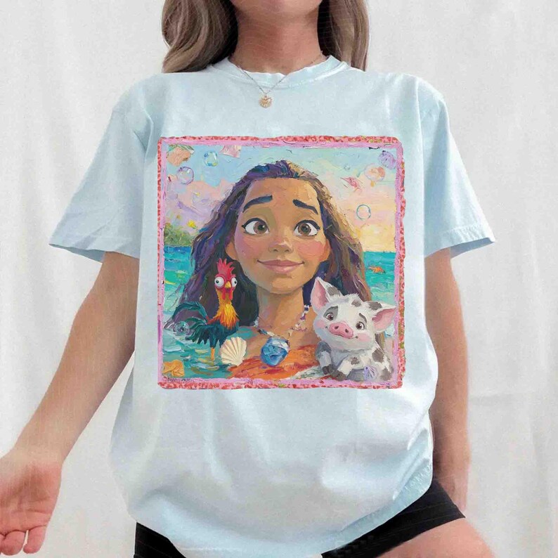 Puede incluir: Una camiseta de manga corta azul claro con un estampado gr&aacute;fico rectangular de una persona joven con cabello largo y oscuro, un gallo y un cerdo peque&ntilde;o. La ilustraci&oacute;n se sit&uacute;a sobre un fondo de oc&eacute;ano con un borde rosa texturizado.