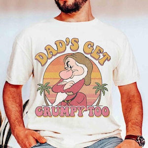 May include: Cream-colored t-shirt featuring a retro design with the text "DAD'S GET GRUMPY TOO." The graphic includes an illustration of Grumpy from Snow White, set against a sunset backdrop with palm trees. The design has a vintage, distressed look.