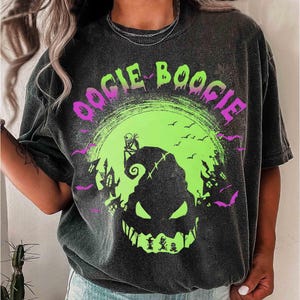 以下が含まれることがあります： 「Oogie Boogie」の文字が入った、ネオングリーンと紫色のグラフィックデザインが施されたダークグレーのTシャツ。不気味な顔、お化け屋敷、コウモリのデザインで、ハロウィンにぴったりです。
