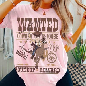 Pode incluir: Camiseta rosa claro com um desenho de um cowboy montando um burro, com o texto "WANTED COWBOY ON THE LOOSE". O design inclui estrelas, um cacto e as palavras "Roundup Reward".