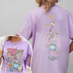 Op de afbeelding: Een lichtpaars t-shirt met afbeeldingen van Toy Story-personages. De voorkant toont Woody, Jessie, Buzz Lightyear en andere figuren met de tekst "TOY story" en "SINCE 1995". De achterkant toont een verticale rangschikking van personages en iconen.