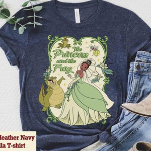 K&ouml;nnte beinhalten: Heather Navy Bella T-Shirt mit farbenfroher Grafik aus "The Princess and the Frog". Das Design zeigt Prinzessin Tiana, einen Frosch und den Text "The Princess and the Frog" in gr&uuml;ner Schrift. Das Shirt wird mit blauen Jeans und einem Paar Stiefel kombiniert.