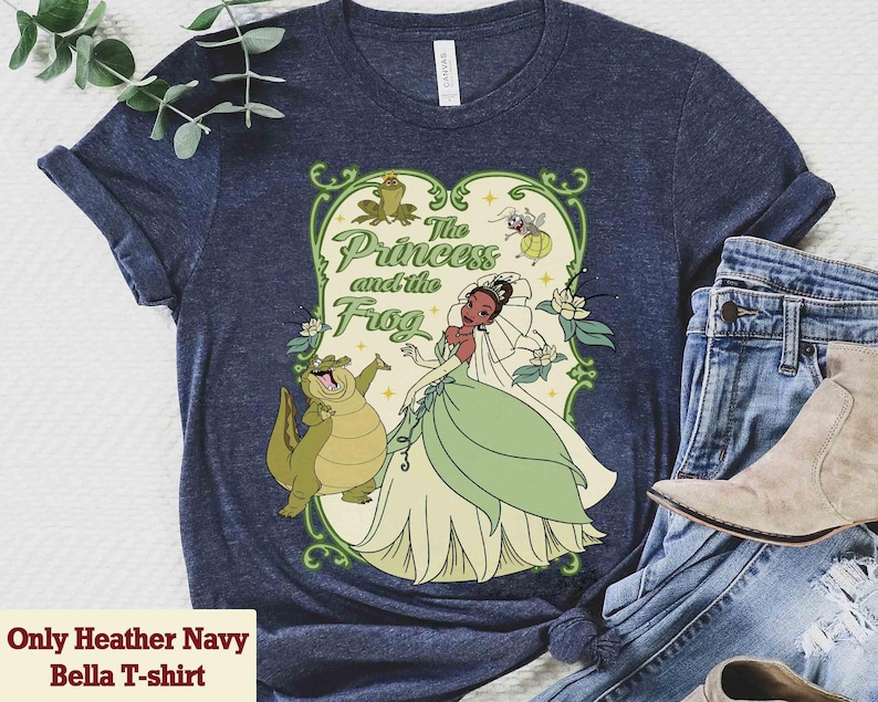K&ouml;nnte beinhalten: Heather Navy Bella T-Shirt mit farbenfroher Grafik aus "The Princess and the Frog". Das Design zeigt Prinzessin Tiana, einen Frosch und den Text "The Princess and the Frog" in gr&uuml;ner Schrift. Das Shirt wird mit blauen Jeans und einem Paar Stiefel kombiniert.