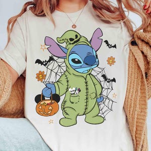 May include: A cream-colored t-shirt featuring a Halloween-themed Stitch character in a green costume holding a pumpkin-shaped candy bucket. The design includes spiderwebs, bats, and flowers. The character has blue skin and purple ears.