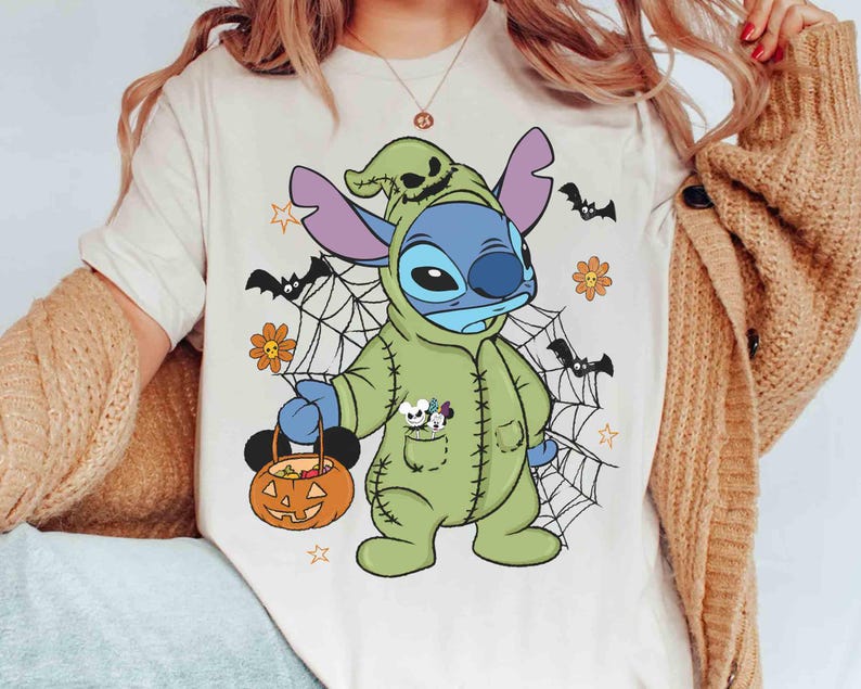 May include: A cream-colored t-shirt featuring a Halloween-themed Stitch character in a green costume holding a pumpkin-shaped candy bucket. The design includes spiderwebs, bats, and flowers. The character has blue skin and purple ears.