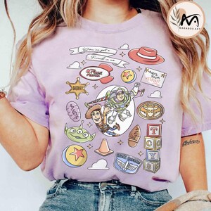 May include: A light purple t-shirt featuring a colorful graphic design with characters and elements from the animated film "Toy Story." The design includes Buzz Lightyear, Woody, and other iconic imagery, with the text "You've got a friend in me."