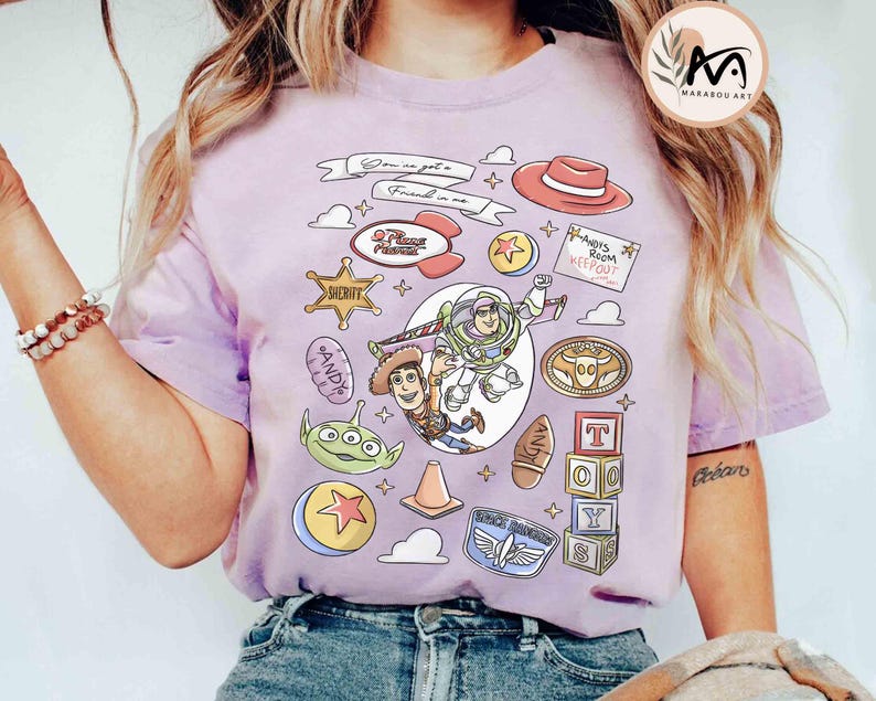 May include: A light purple t-shirt featuring a colorful graphic design with characters and elements from the animated film "Toy Story." The design includes Buzz Lightyear, Woody, and other iconic imagery, with the text "You've got a friend in me."