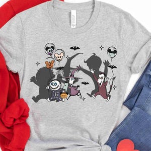 Puede incluir: Una camiseta gris claro con un dise&ntilde;o de dibujos animados de Halloween. El gr&aacute;fico representa varios personajes, incluyendo un esqueleto, una bruja y un vampiro, con globos y murci&eacute;lagos. La camiseta est&aacute; hecha de un material suave.