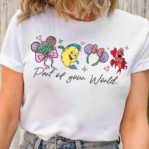 Puede incluir: Camiseta blanca con ilustraciones de dibujos animados de personajes de una pel&iacute;cula animada popular. El dise&ntilde;o incluye una cola de sirena, un pez amarillo, un par de orejas de rat&oacute;n y un cangrejo rojo. El texto "Part of your World" est&aacute; escrito debajo.