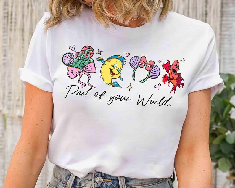 Puede incluir: Camiseta blanca con ilustraciones de dibujos animados de personajes de una pel&iacute;cula animada popular. El dise&ntilde;o incluye una cola de sirena, un pez amarillo, un par de orejas de rat&oacute;n y un cangrejo rojo. El texto "Part of your World" est&aacute; escrito debajo.
