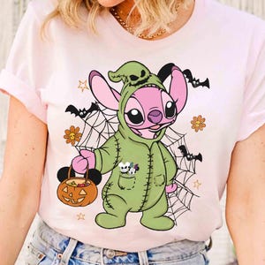 May include: Pink t-shirt featuring a cartoon character in a green Halloween costume holding a pumpkin-shaped candy bucket. The design includes bats, spiderwebs, and flowers, creating a festive Halloween theme.