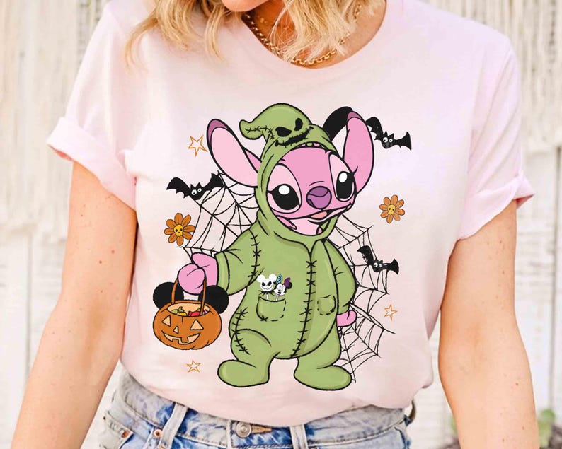 May include: Pink t-shirt featuring a cartoon character in a green Halloween costume holding a pumpkin-shaped candy bucket. The design includes bats, spiderwebs, and flowers, creating a festive Halloween theme.