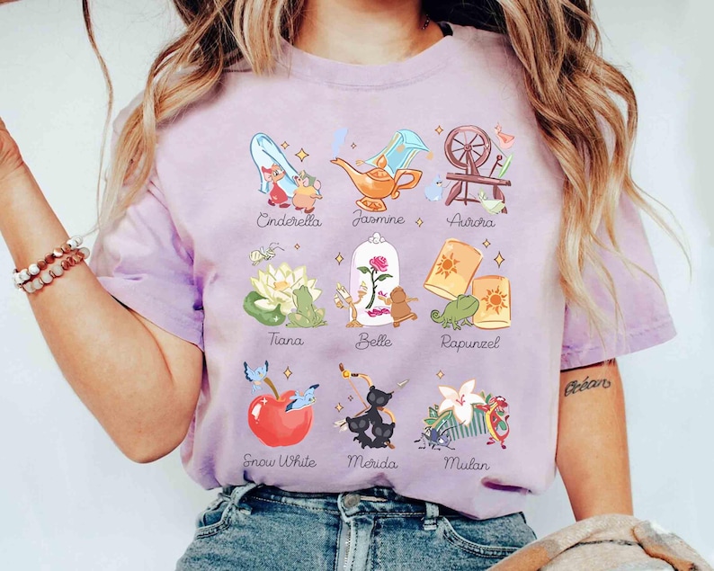 May include: A light purple t-shirt featuring illustrations of various Disney princesses and their associated icons. Cinderella's glass slipper, Jasmine's lamp, and Aurora's spinning wheel are among the images. The names of the princesses are written below each image.