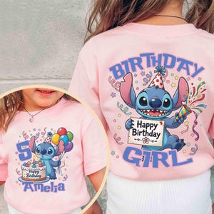 May include: Pink short-sleeved t-shirt featuring a cartoon character holding balloons and a birthday cake. The shirt has the text "Happy Birthday" and the name "Amelia". The back of the shirt says "Birthday Girl".