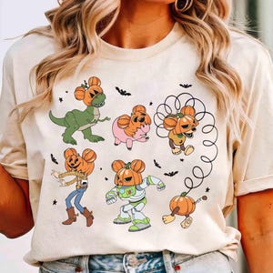 May include: Beige t-shirt featuring cartoon characters with pumpkin heads. The characters include a dinosaur, a pig, a dog, a cowboy, a space ranger, and a pumpkin on wheels. Black bats and stars are scattered around the characters.