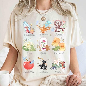 May include: Cream-colored t-shirt featuring colorful illustrations of Disney princesses and their associated icons. The shirt includes images of Cinderella, Jasmine, Aurora, Tiana, Belle, Rapunzel, Snow White, Merida, and Mulan, with their names below.