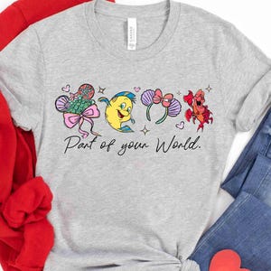 Puede incluir: Camiseta gris claro con personajes de dibujos animados coloridos y el texto "Part of your World". El dise&ntilde;o incluye una cola de sirena, un pez amarillo, un par de orejas moradas con un lazo y un cangrejo rojo.