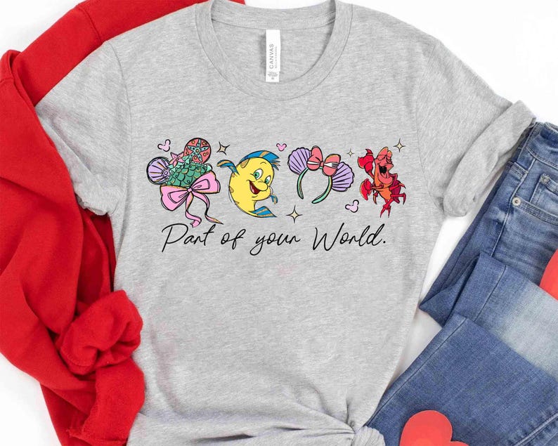 Puede incluir: Camiseta gris claro con personajes de dibujos animados coloridos y el texto "Part of your World". El dise&ntilde;o incluye una cola de sirena, un pez amarillo, un par de orejas moradas con un lazo y un cangrejo rojo.