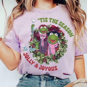 Puede incluir: Camiseta lila claro con un diseño navideño. Presenta dos personajes de Kermit la Rana con ropa de invierno, enmarcados por una corona. El texto dice "'Tis the Season to be Jolly & Joyous."
