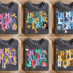 Op de afbeelding: Zes donkergrijze t-shirts met kleurrijke retro-ontwerpen. Elk shirt heeft de zin "In My" gevolgd door de naam van een prinses en "Era". De ontwerpen bevatten afbeeldingen van Aurora, Jasmine, Elsa, Rapunzel, Ariel en Belle, met bijbehorende iconen.